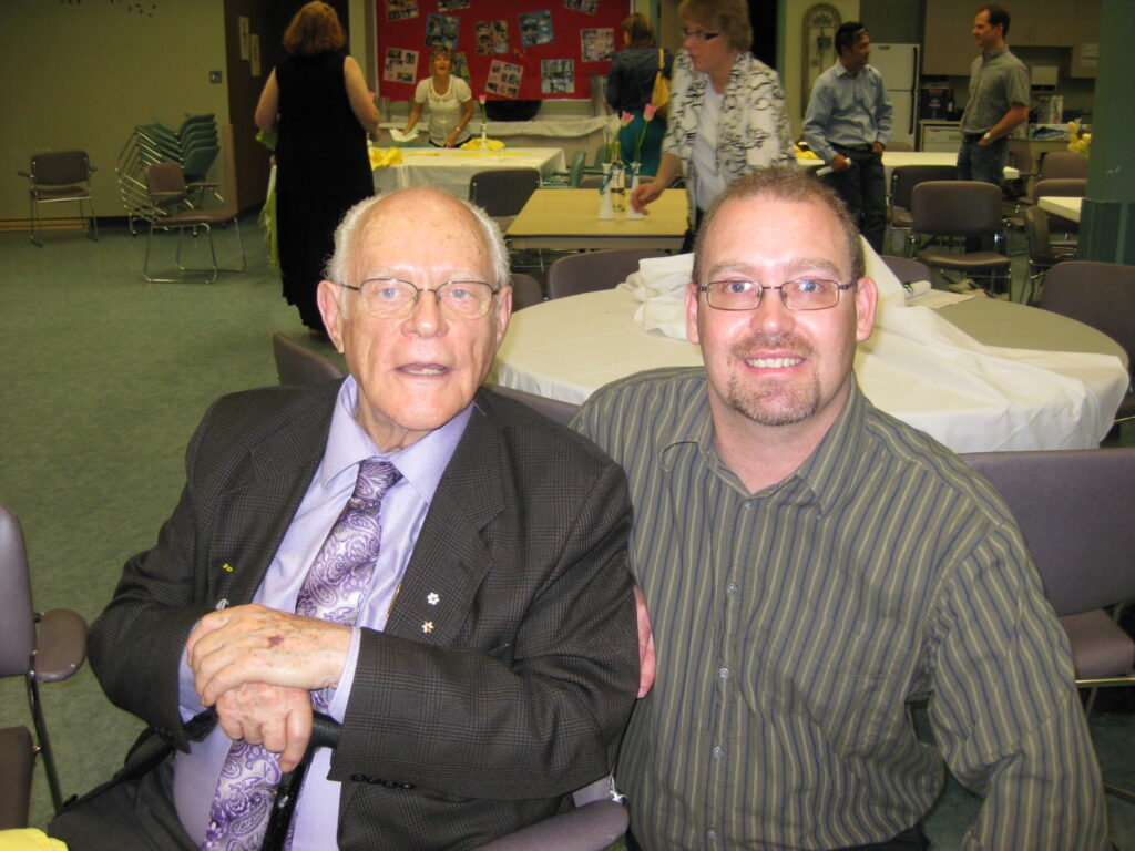 a man with white hair, a purple shirt and tie and black suit jacket, sitting with a man with a striped shirt