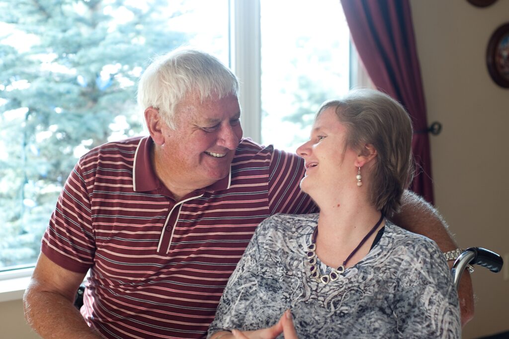 a smiling, white haired man in a red striped golf shirt with his arm around a smiling woman in a floral white and black top who is sitting in a wheelchair