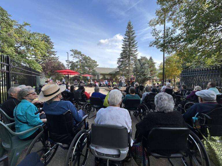 A crowd of over 80 people of all ages, seated in a circle in wheelchairs and chairs, participating in an outdoor concert. 4 singers are playing guitar and the area is surrounded by green trees, blue sky, and fencing.